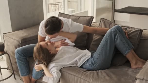 A positive lovely young couple man and woman in casual wear are kissing sitting on the sofa together in the living room - Filmati, video