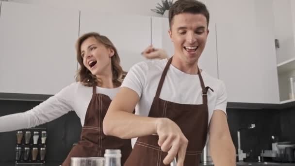 Positive smiling young couple man and woman wearing brown aprons are making dough while dancing in the kitchen - 映像、動画