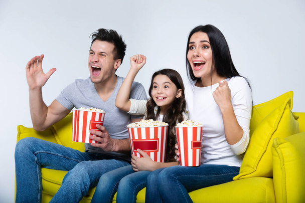 Happy young family watching tv at home with popcorn sitting on yellow sofa - Φωτογραφία, εικόνα
