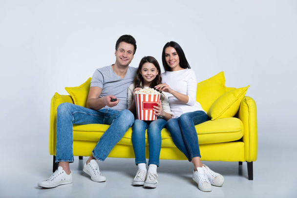 Happy young family watching tv at home with popcorn sitting on yellow sofa - Фото, изображение