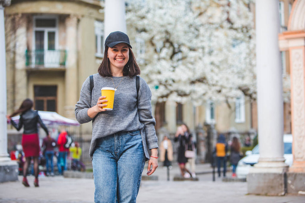 若いスタイリッシュな女性がコーヒーカップで通りを歩く。春咲きの木を背景に - 写真・画像