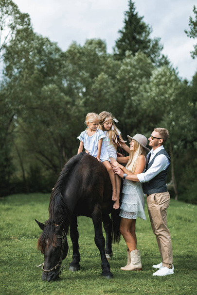 Young stylish family in cowboy wear, father, mother teaching their two little daughters to ride a horse. Family horse riding on the nature in countryside - Foto, afbeelding