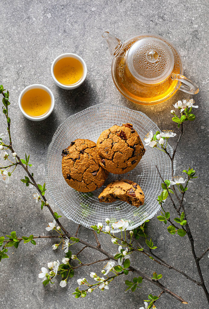 homemade oatmeal cookies on a table with branches of a blossoming tree and green tea in a glass teapot. - Photo, Image