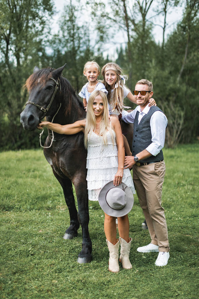 Cute two happy blond girls children riding horse on nature trees background, having walk with young parents, looking at camera and smiling. Stylish young family walking with brown horse in the meadow - Foto, afbeelding