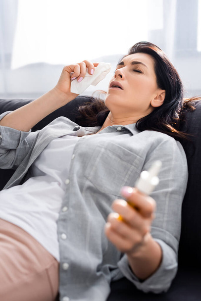 selective focus of allergic woman holding napkin and nasal spray while lying on sofa  - Photo, Image
