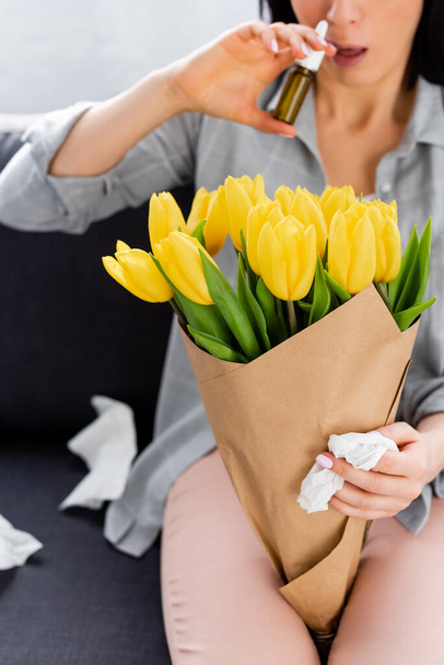 widok upraw kobiety z alergią na pyłki siedząc na kanapie z kwiatami i trzymając aerozol do nosa  - Zdjęcie, obraz