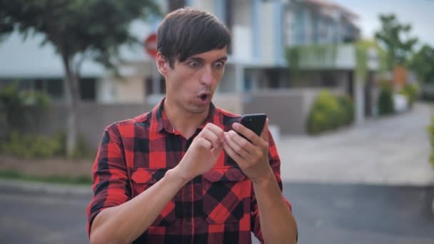 Scared Young Man está sorprendido por lo que vio en su teléfono inteligente, de pie fuera en la calle de la ciudad. Hombre sorprendido lee malas noticias en su Smartphone
. - Imágenes, Vídeo