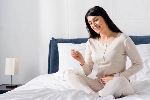 happy woman holding pregnancy test and sitting on bed  - Foto, Imagem