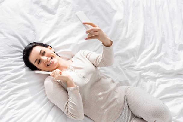 top view of happy woman taking selfie with pregnancy test while lying on bed  - Foto, imagen