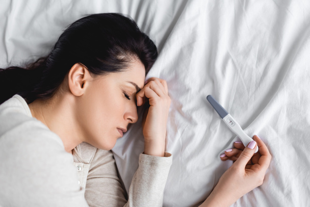 top zicht van depressieve vrouw met gesloten ogen liggend op bed in de buurt van zwangerschapstest  - Foto, afbeelding