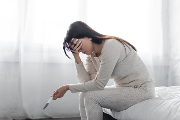 Traurige Frau sitzt auf Bett und hält Schwangerschaftstest mit negativem Ergebnis  - Foto, Bild
