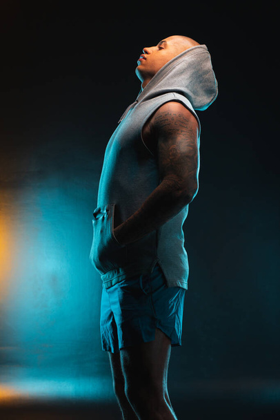 Young attractive man wearing a vest with a hood on his head. Orange and teal light on the background - Фото, зображення