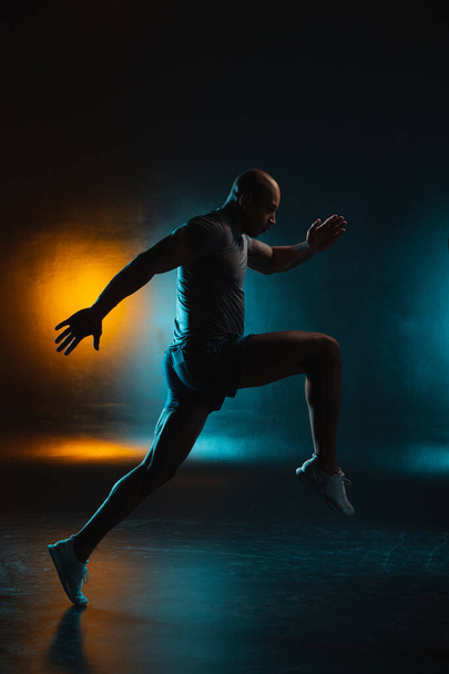 Young bold sportsman moving on teal orange background. Running pose - Foto, afbeelding