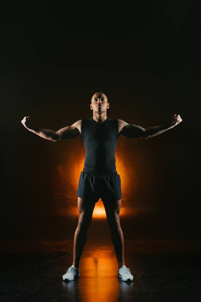 Handsome strong sportsman in sporty clothes showing biceps. Orange light on the background - Фото, зображення