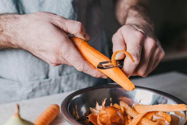 Rough male hands clean a carrot with a special knife on the background of the kitchen - rustic style - Fotoğraf, Görsel