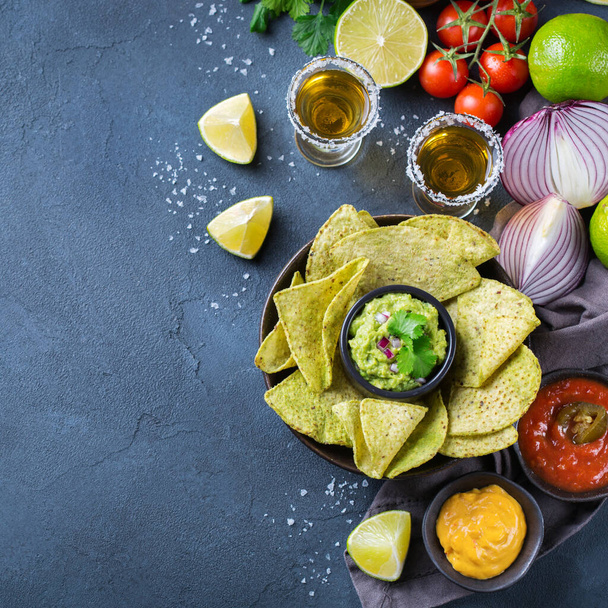 Yellow corn tortilla chips nachos with guacamole, red hot jalapeno chili salsa and cheese sauce with tequila on a dark table. Top view flat lay background  - Foto, Imagem