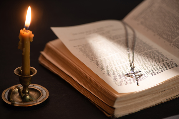 KYIV, UKRAINE - JANUARY 17, 2020: church candle in candlestick burning near bible with catholic cross in dark with sunlight - Foto, afbeelding