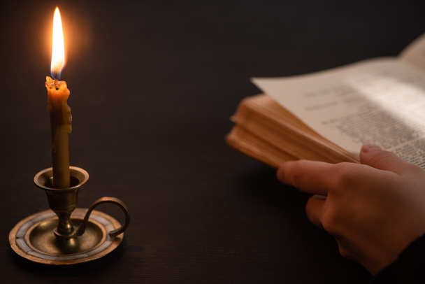 cropped view of woman holding holy bible near burning candle in dark - Photo, Image