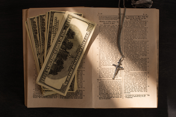 KYIV, UKRAINE - JANUARY 17, 2020: top view of holy bible with money and cross on dark background with sunlight - Fotografie, Obrázek