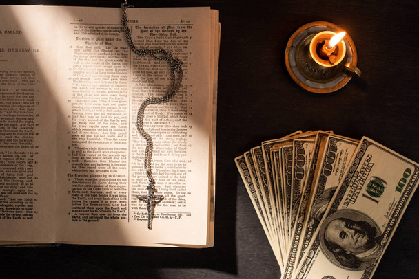 KYIV, UKRAINE - JANUARY 17, 2020: top view of holy bible with money, cross and candle on dark background with sunlight - Φωτογραφία, εικόνα