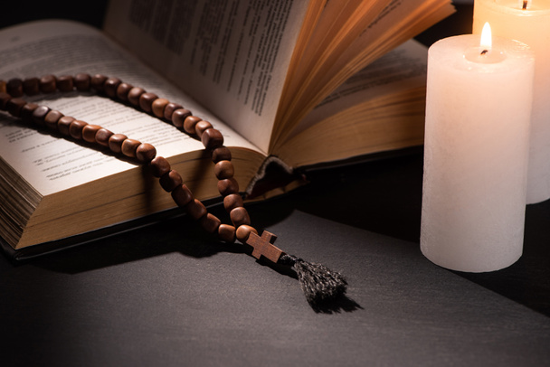 holy bible with rosary on black dark background with burning candles - Фото, изображение
