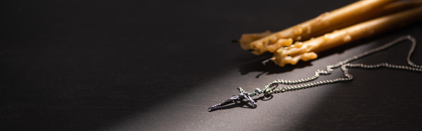 kříž a kostelní svíčky na černém tmavém pozadí se slunečním světlem, panoramatický záběr - Fotografie, Obrázek