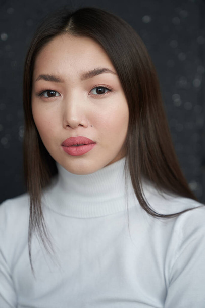  A portrait of a young Asian girl holding her hands to her face looks directly at the camera. She's wearing a light turtleneck. The concept of skin care and hair                                    - Fotoğraf, Görsel