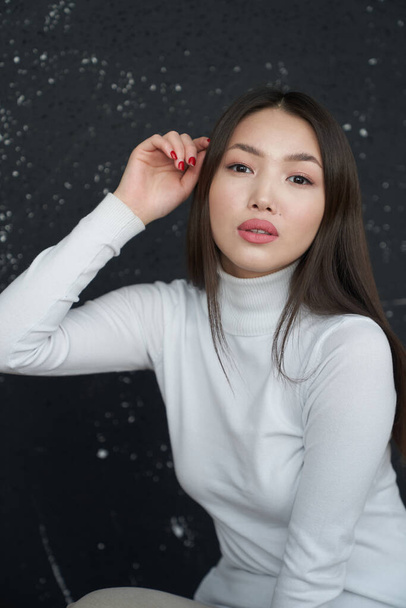 Portrait of a young Asian girl holding her hands to her face, looking directly at the camera. She's wearing a light turtleneck. The concept of skin care and hair                                      - Фото, изображение