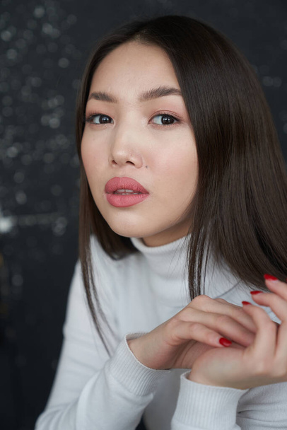  Portrait of a young Asian girl holding her hands to her face, looking directly at the camera. She's wearing a light turtleneck. The concept of skin care and hair                                      - Photo, Image