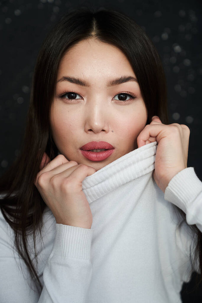 Portrait of a young Asian girl with the collar turned up from her turtleneck to her face, looking directly at the camera, in a light jumper. The concept of skin care and hair                                - Фото, зображення