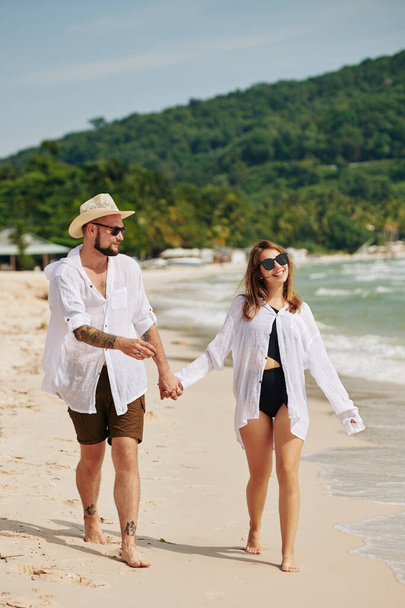Gelukkige jonge man en vrouw in een zonnebril en witte shirts hand in hand op het strand lopen - Foto, afbeelding