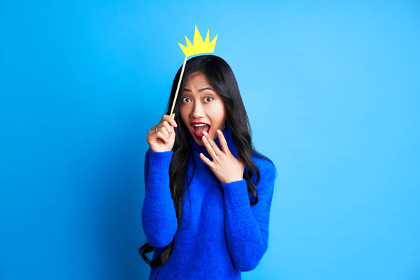 Surprised beautiful woman hold a paper craft crown on a stick on blue background. Party, emotions concept - Фото, зображення