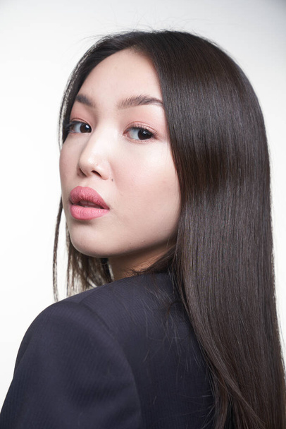 Portrait of a young beautiful Asian girl looking over her shoulder, directly into the camera, in a blue jacket. The concept of skin care and hair                                - Photo, image