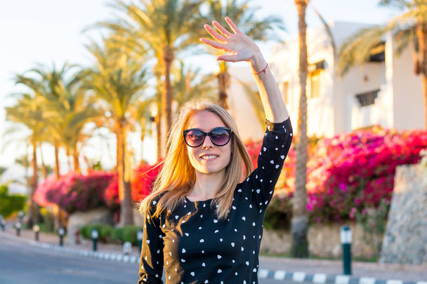 Jovem mulher feliz em óculos de sol acena sua mão na frente de arbustos de hibisco vermelho e rosa florescendo e palmeiras ao pôr do sol
 - Foto, Imagem