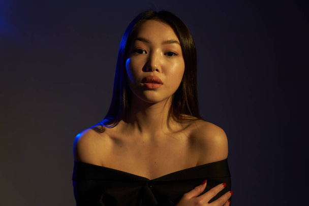 Portrait of a young beautiful Asian girl looking directly at the camera. The woman's hair and shoulders in colorful bright UV blue lights. On black background.                                   - Fotografie, Obrázek