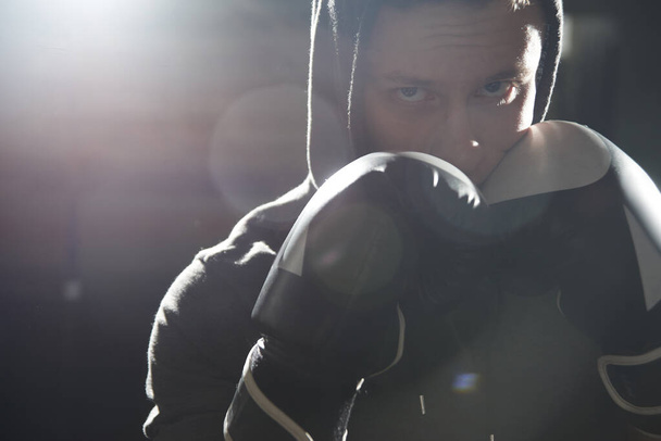 Closeup portrait of a cool professional boxer in gloves and a hood, aggressively looking at the camera, beating with his fist - Photo, Image
