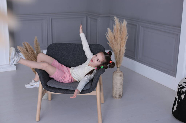  Yedi yaşında genç ve güzel bir kızın romantik portresi. Komik bir saç stili var. Büyük gri bir koltukta oturuyor. - Fotoğraf, Görsel