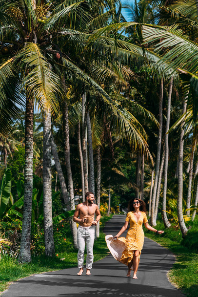 Beautiful couple running among palm trees in Bali, Indonesia. Honeymoon on the Islands. A happy couple in love runs along the road. Honeymoon trip. Wedding trip. Couple on vacation in Bali - Foto, afbeelding
