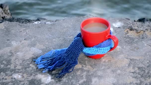 Red mug with coffee and steam tied with blue knitted scarf stands on stone - Materiał filmowy, wideo