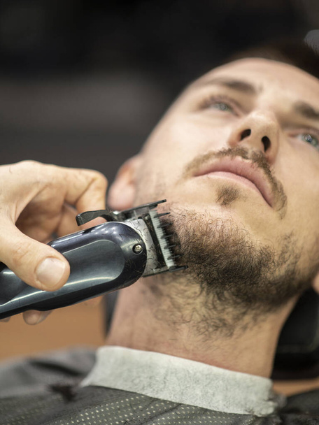 Barber using electric razor while trimming a beard of male customer at barber shop - Valokuva, kuva