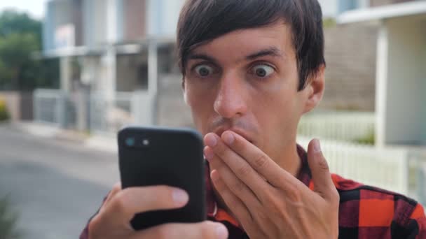 Portrét vyděšeného mladíka je šokován tím, co viděl na svém chytrém telefonu, jak stojí venku na ulici. Šokovaný muž čte špatné zprávy na svém smartphonu. - Záběry, video