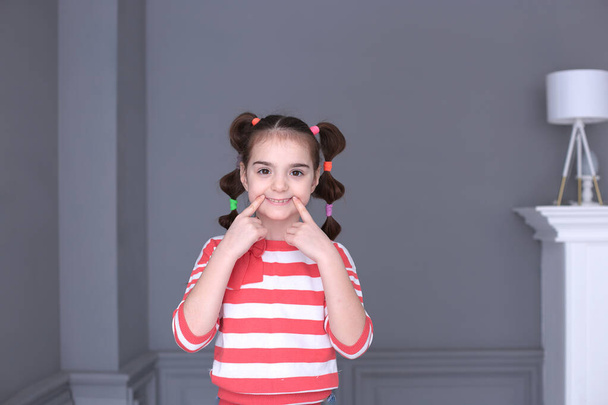 portrait of a young funny girl of seven years old with a funny hairstyle with a striped sweater in a good mood and grimaces on a gray background - Photo, Image