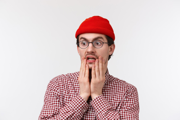 Oh gosh omg. Surprised and speechless hipster guy with beard in red beanie, glasses, gasping staring at something shocking gross, look left spot terrible thing, standing white background - Photo, Image