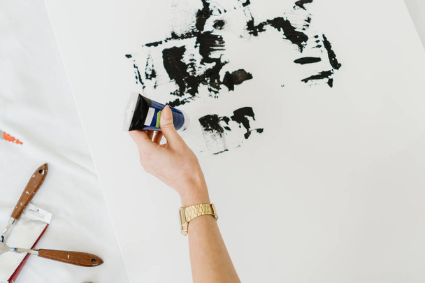 Рука жінки малює на білому полотні, використовуючи чорні кольори олії. Багато інструментів для малювання біля підлоги
. - Фото, зображення