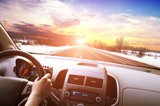 Car dashboard with driver hand on the black steering wheel against winter asphalt road and sky with sunset - Fotó, kép