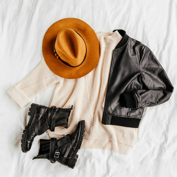 Leather jacket and white warm sweater with brown hat and boots on white bed. Women's stylish autumn or winter outfit. Trendy clothes collage. Flat lay, top view.  - Foto, imagen