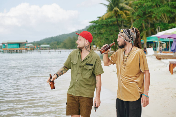 Amici maschi che camminano sulla spiaggia, bevono birra in bottiglia e si godono il paesaggio marino - Foto, immagini