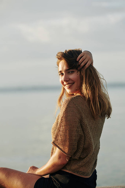 Gün batımında sahilde oturup kameraya dönen sevimli, çekici genç bir kadın. - Fotoğraf, Görsel