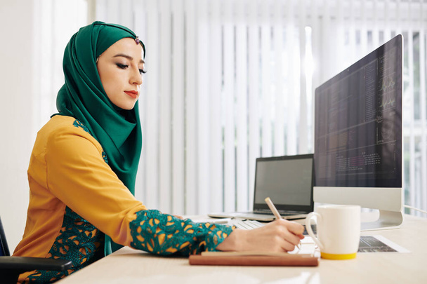 Jeune femme musulmane prenant des notes dans le planificateur lorsque vous travaillez sur le code de programmation pour un nouveau projet
 - Photo, image