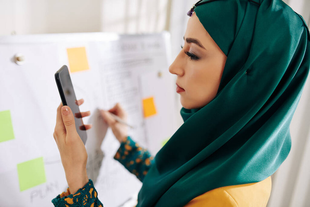 Kaunis muslimi projektipäällikkö tarkistaa tiedot älypuhelimen näytöllä, kun kirjoitat taululle ja valmistaudut esittelyyn
 - Valokuva, kuva